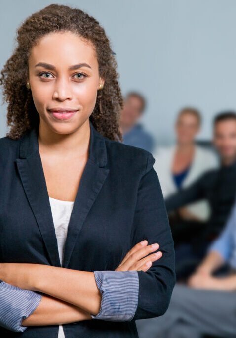 Portrait of female African American executive manager standing with crossed arms in meeting room, business people sitting at office desk on background